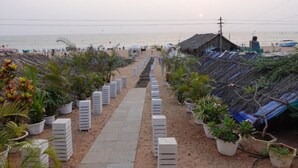 A beachside area with a pathway leading to a beach, surrounded by potted plants and thatched-roof structures. - Lillywoods Zac Beach Resort (Calangute)