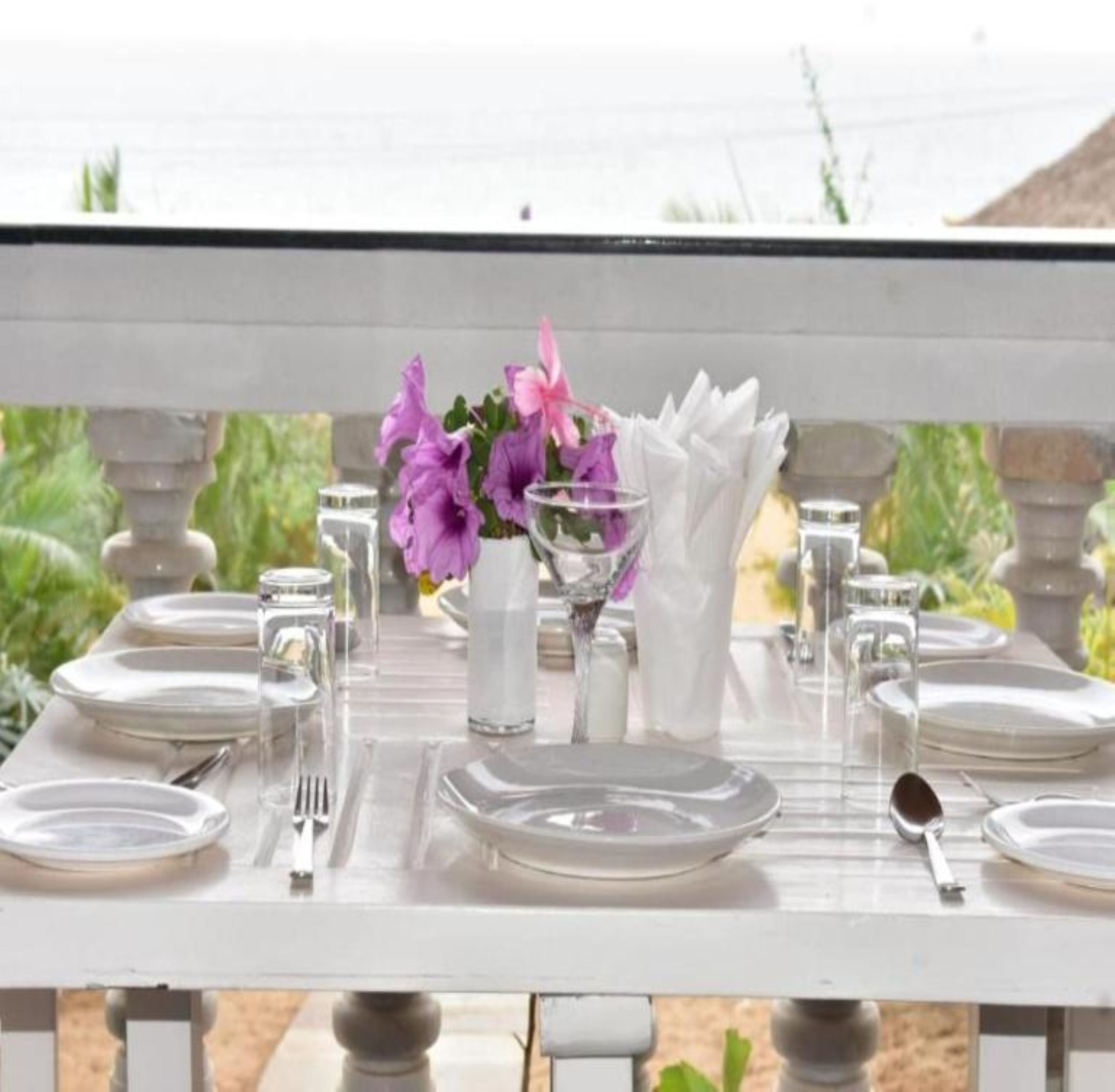 Mesa posta para refeição com pratos brancos, talheres e um vaso de flores roxas.