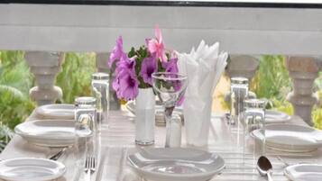 Mesa posta para refeição com pratos brancos, talheres e um vaso de flores roxas.