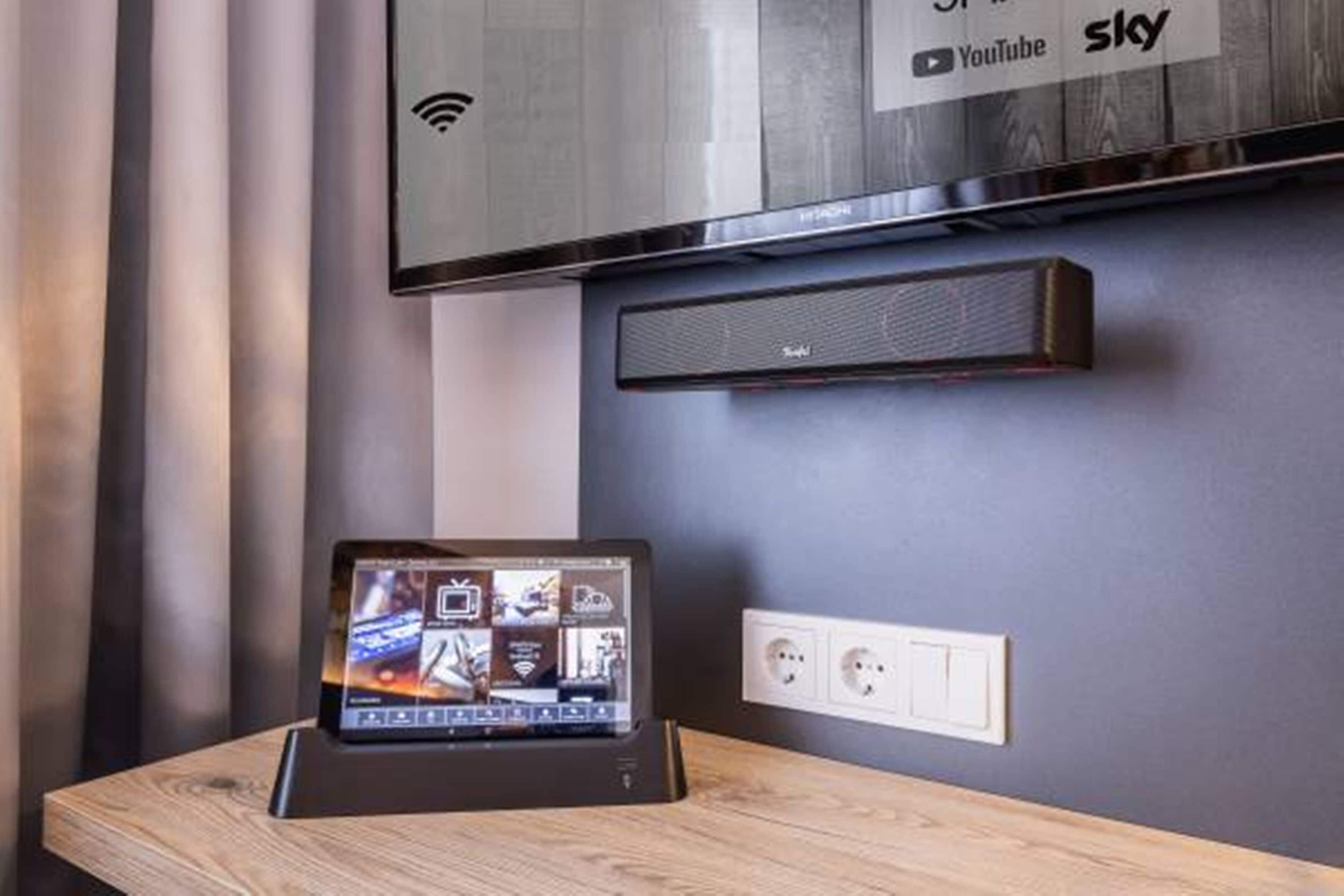 Una habitación de hotel moderna con un televisor de pantalla plana en la pared, una pequeña mesa con una tableta mostrando un menú de servicio a la habitación, y un altavoz en la pared.