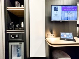 A hotel room with a TV displaying a welcome screen, a coffee machine, a microwave, and a small table with a laptop.