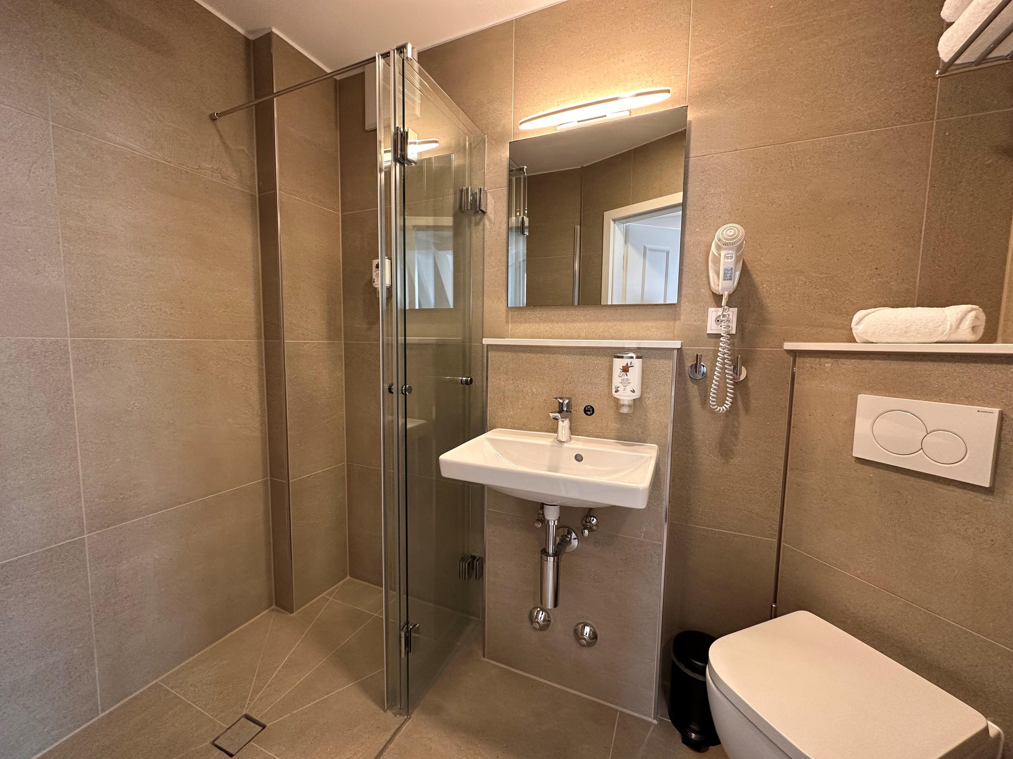 Un baño moderno con ducha de vidrio, un lavabo blanco, un espejo, un perchero para toallas y un inodoro.