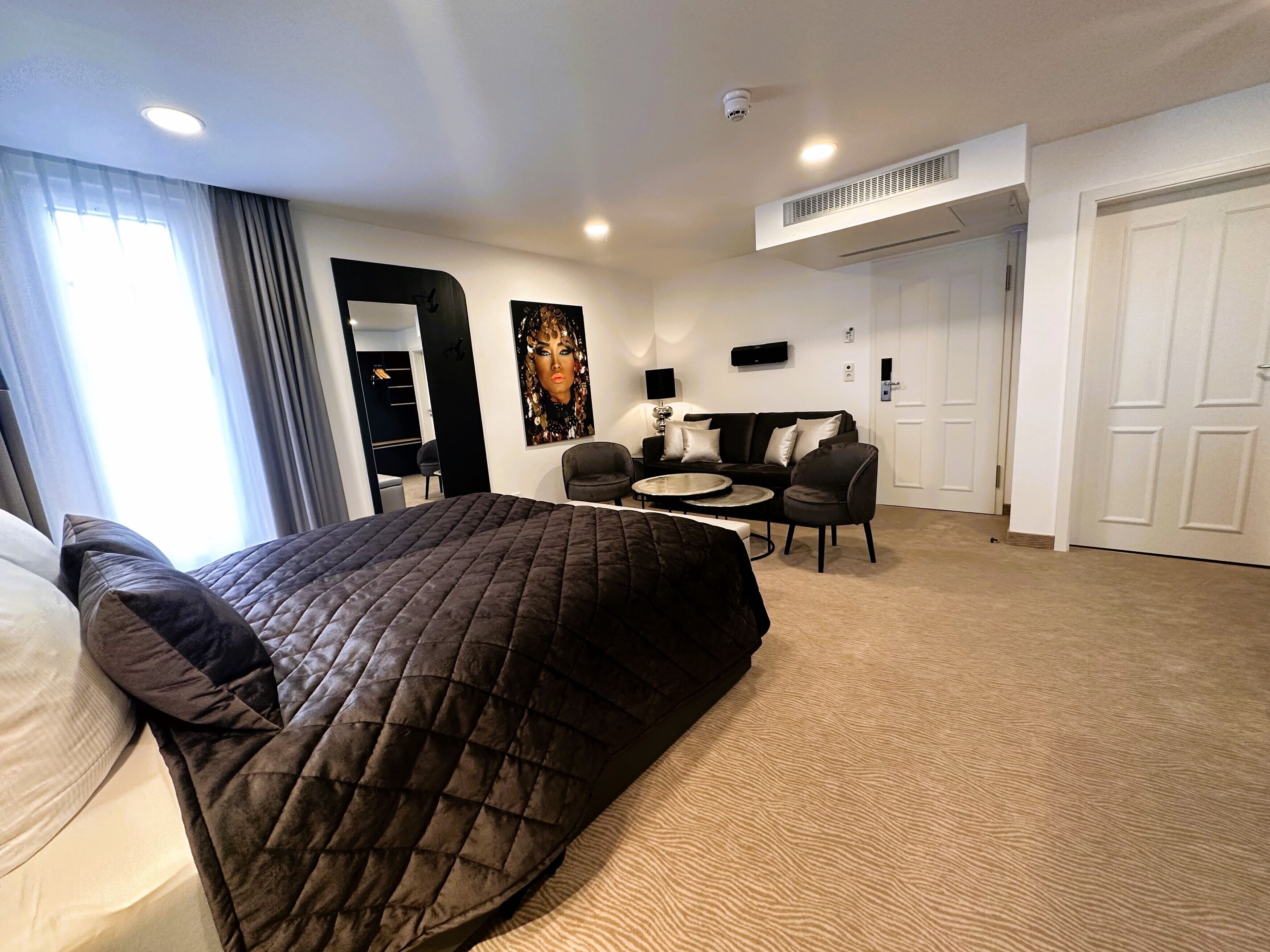 A hotel room with a large bed, a desk, two chairs, a small table, a painting on the wall, and a door.