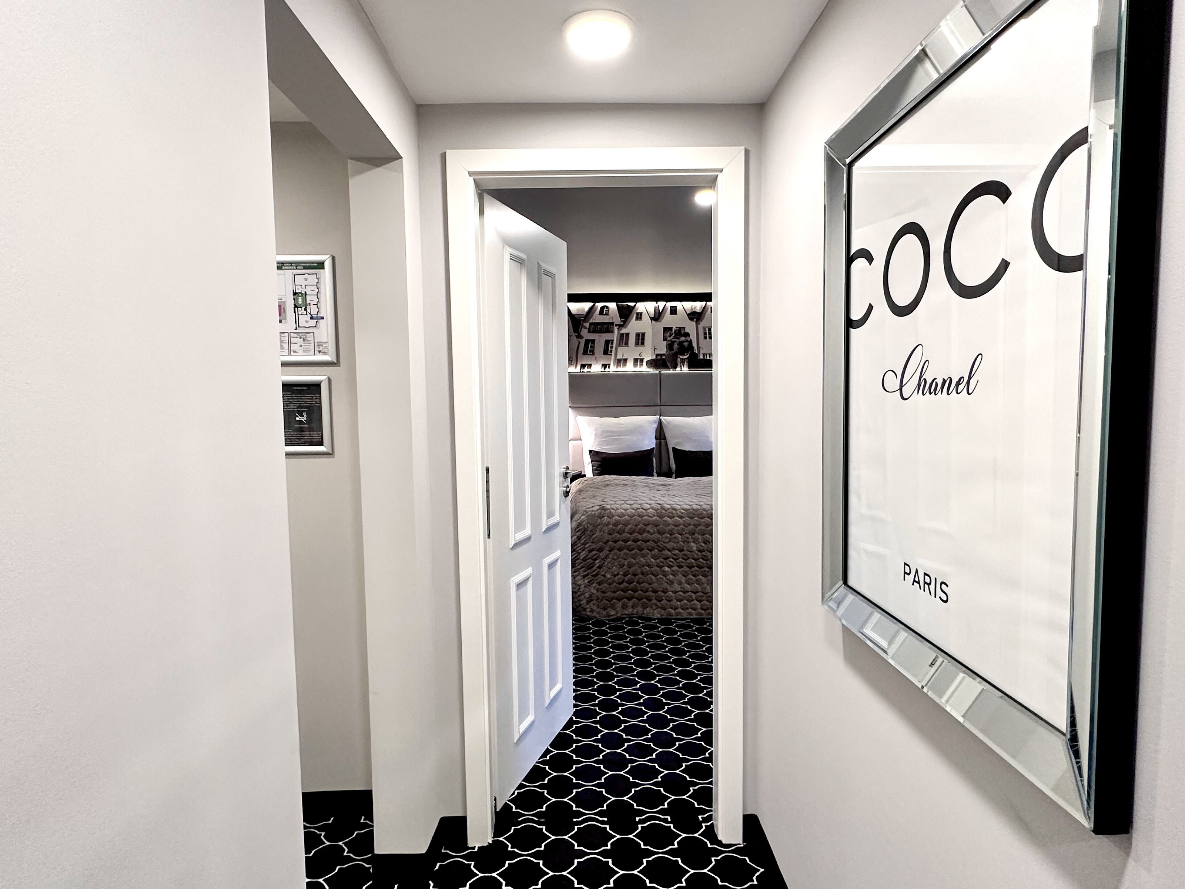 Käytävän lattia on mustavalkoista kuvioitua parkettia, siellä on ovi, joka johtaa makuuhuoneeseen, ja seinällä on kehystetty kyltti, jossa lukee »COCO Chanel Paris».