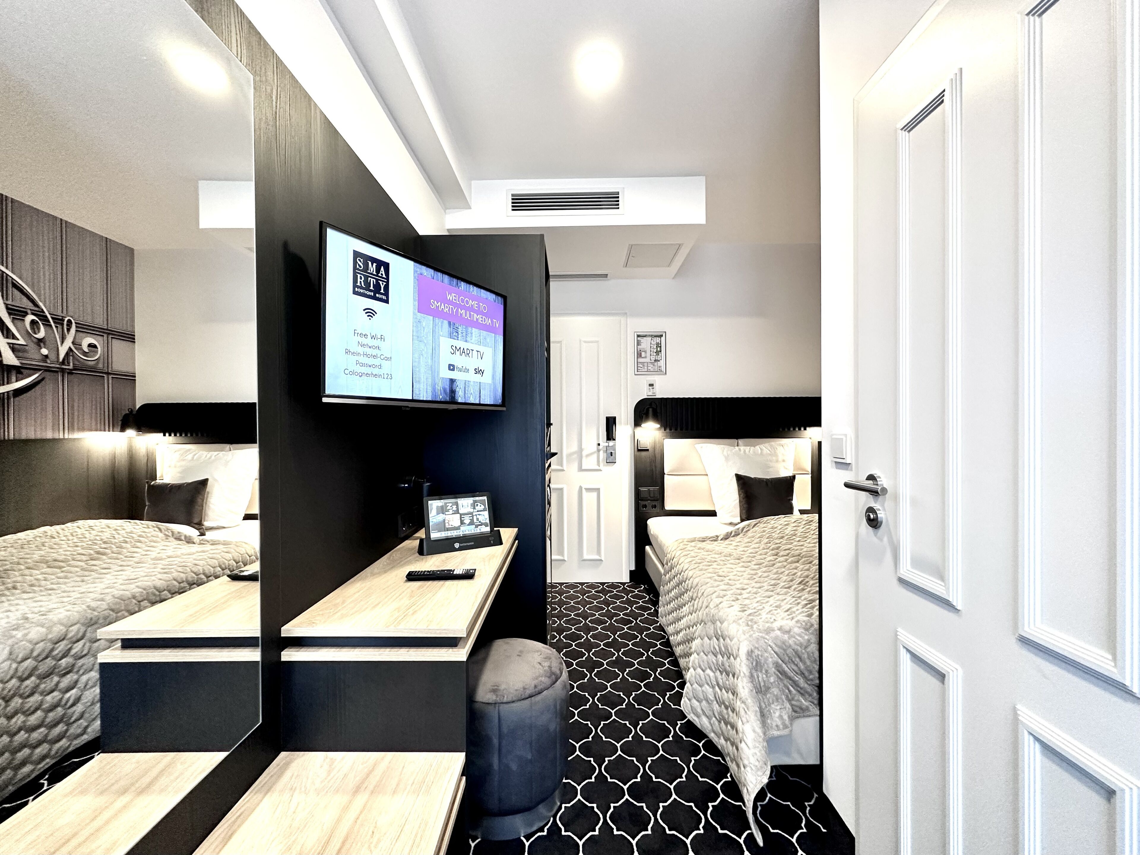 A modern hotel room with two beds, a TV displaying welcome messages, a desk with a laptop, and a patterned floor.
