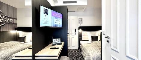 A modern hotel room with two beds, a TV displaying welcome messages, a desk with a laptop, and a patterned floor.