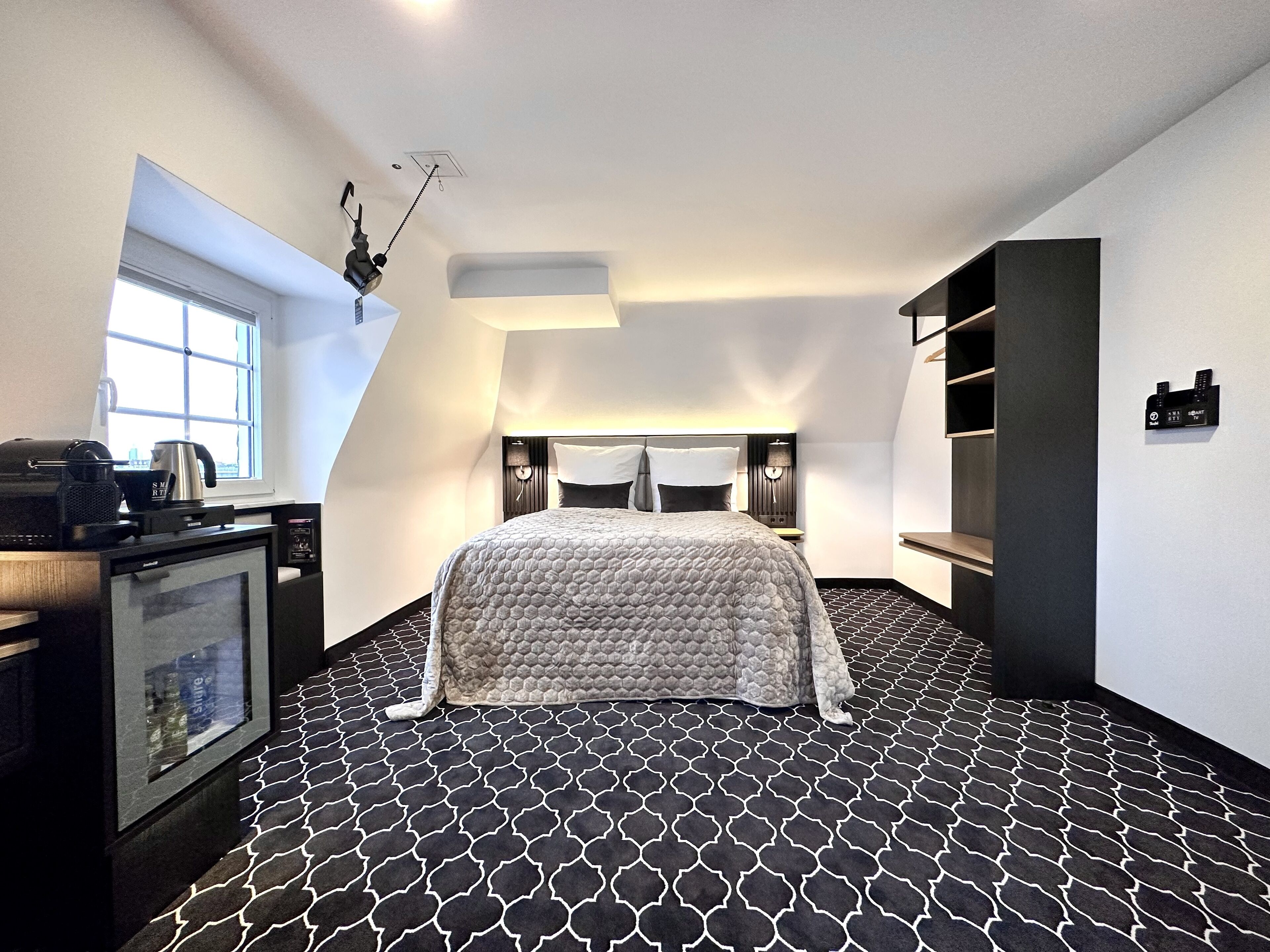 Un dormitorio moderno con piso de diseño geométrico, cama con cabecera acolchada, escritorio con cafetera y televisor montado en la pared.