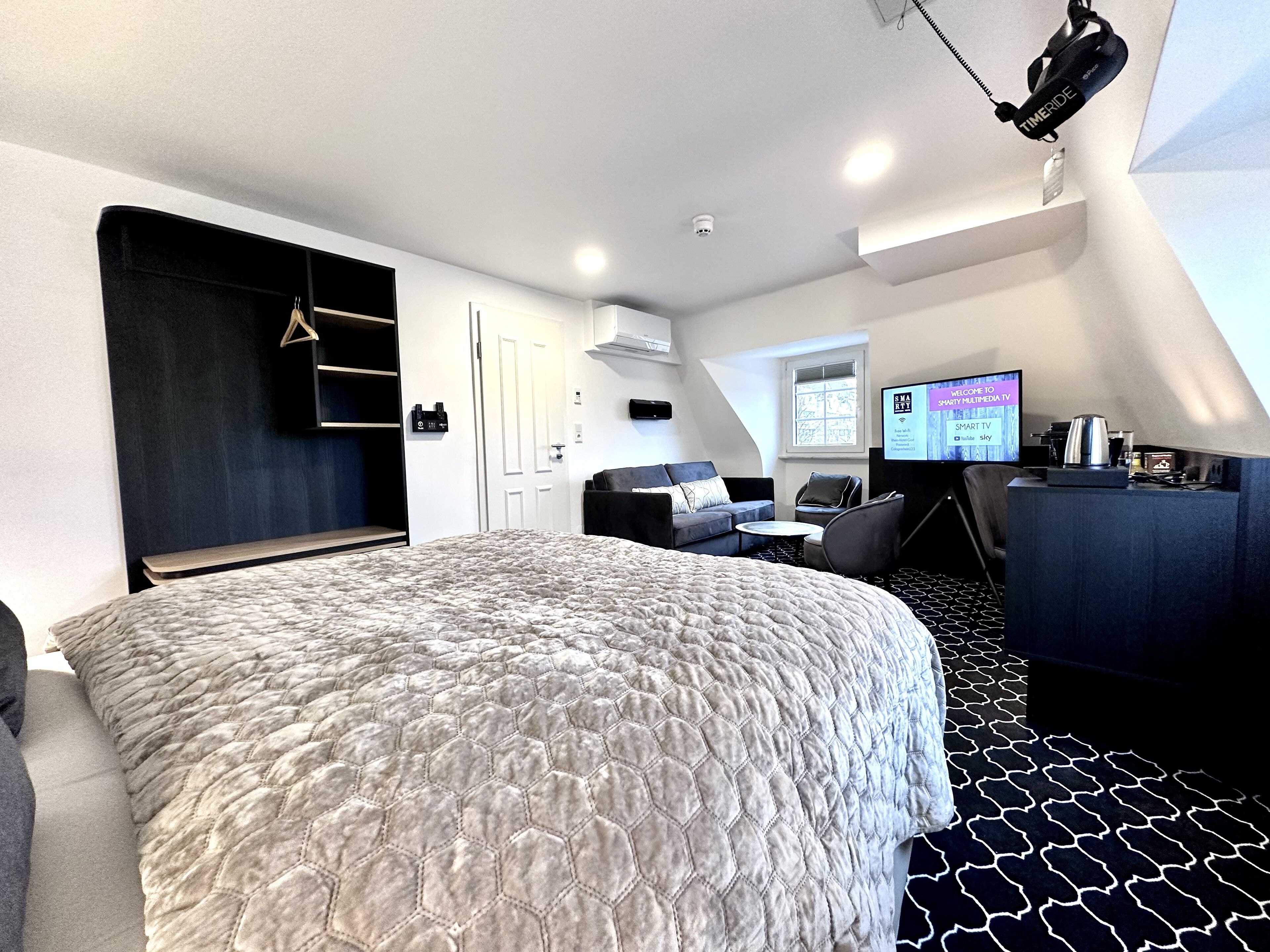 A modern hotel room with a large bed, a desk, a chair, a TV, and a wardrobe.