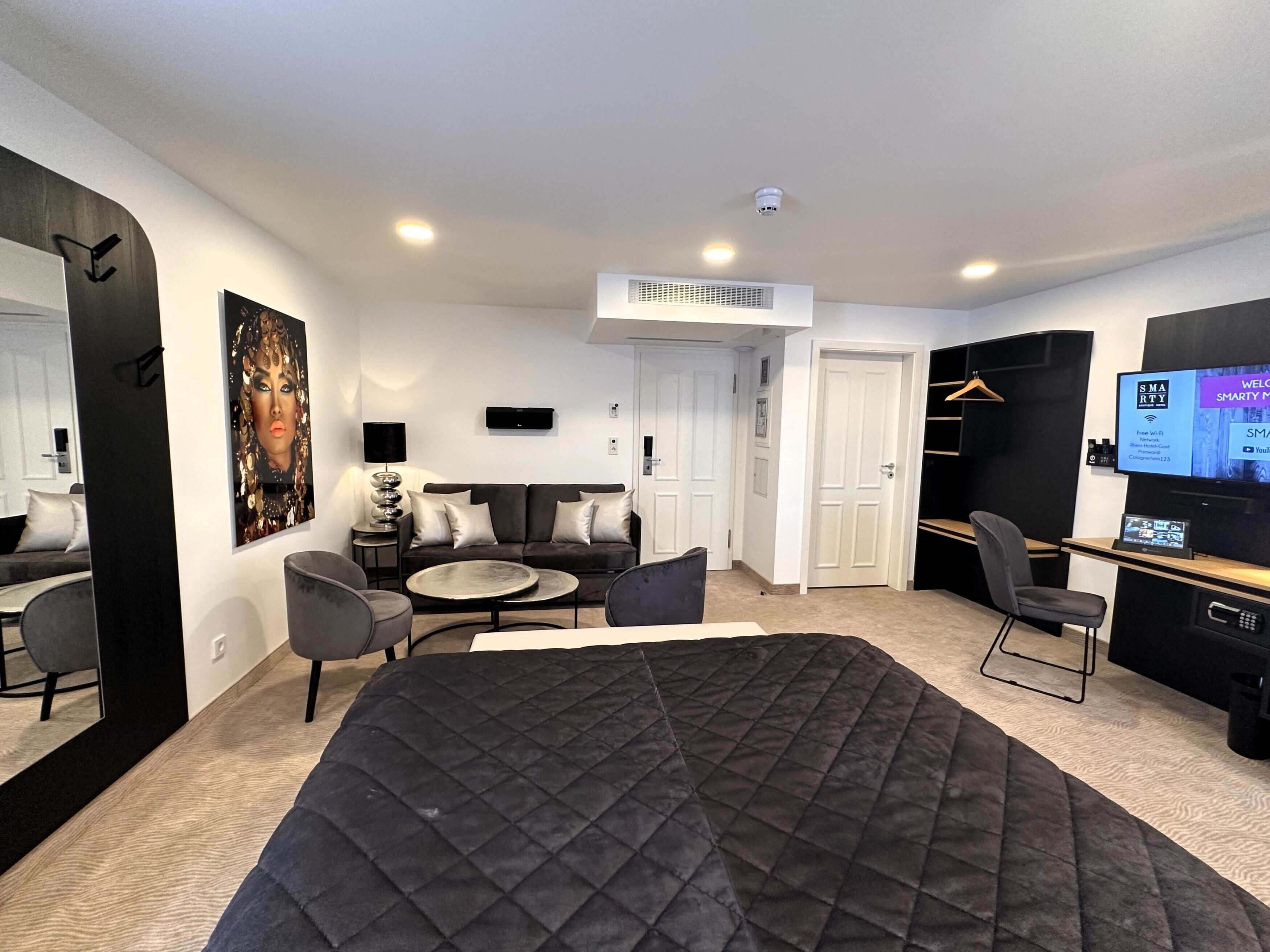 A modern hotel room with a large bed, a sofa, a small table, and a TV. There is a wardrobe and a mirror on one wall.