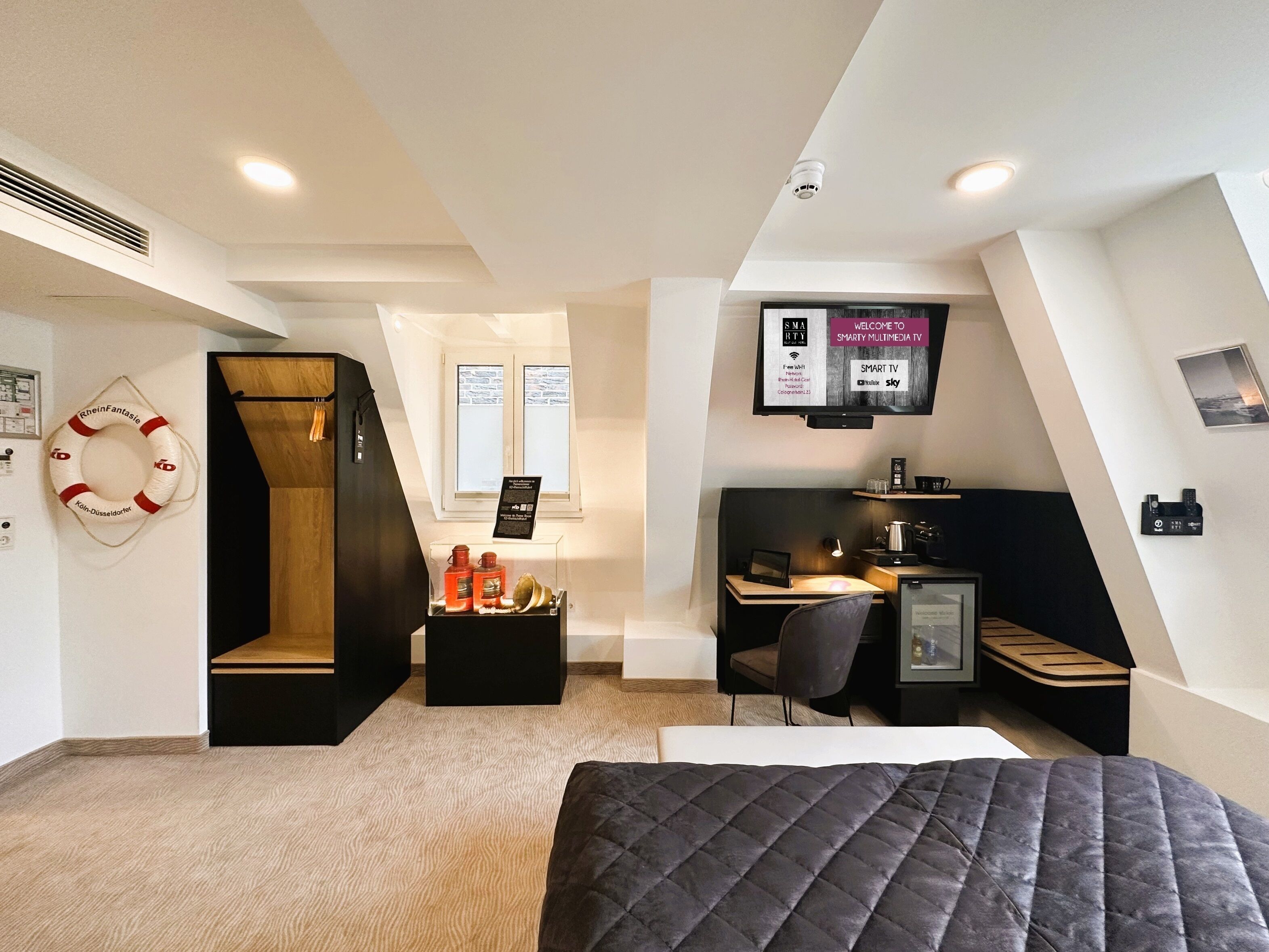 Een moderne hotelkamer met een hoogslaper, een bureau met stoel, een aan de muur bevestigde televisie en een kleine kitchenette.