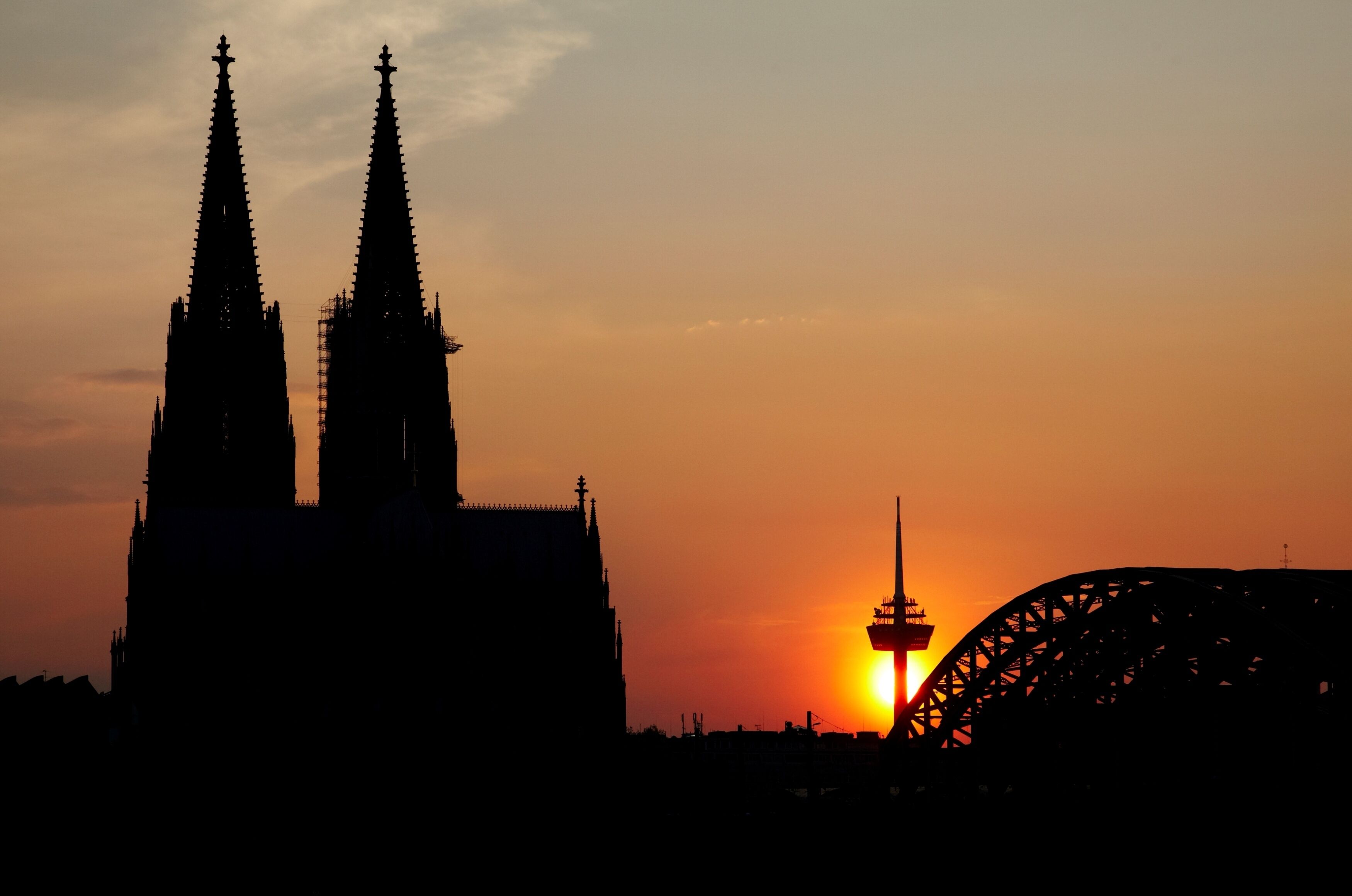 A silhouette of a cathedral with two spires and a bridge with a tower in the background during sunset.