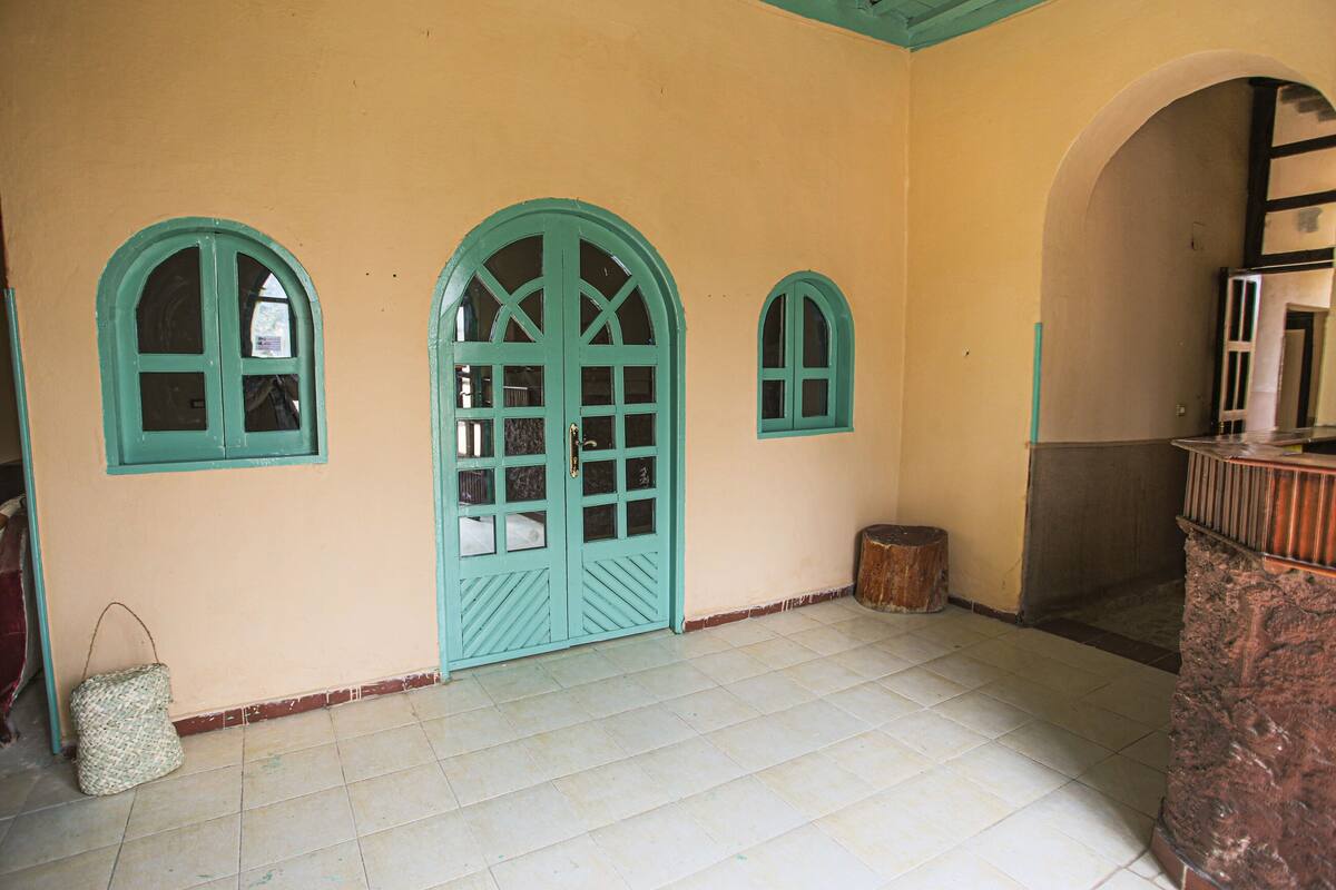 A room with three arched windows and a large door, a tiled floor, and a small wooden stool.