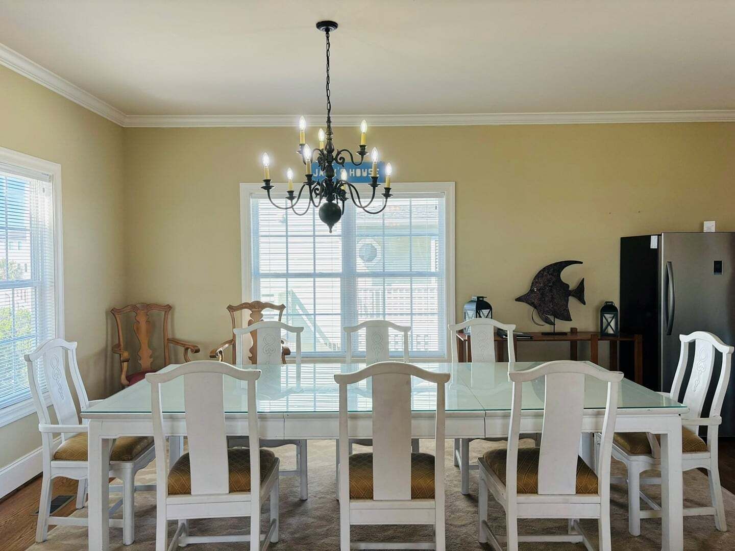 A dining room with a glass table, white chairs, a chandelier, and a kitchen area with a refrigerator.