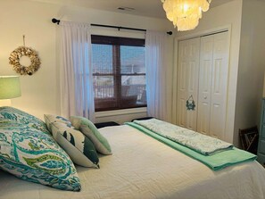A bedroom with a bed, a window with blinds, a door, and a wall-mounted light fixture.