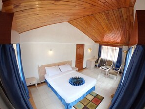 A bedroom with a wooden ceiling, a bed with a blue and white bedspread, a bedside table, a chair, a small table with a lamp, and a window with curtains. - Stay.Plus Hurlingham B&B Hotel (Nairobi)