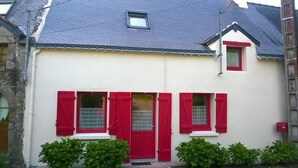 Een huis met twee verdiepingen, een rode deur en luiken, en een klein raam op de bovenverdieping, met een witte gevel.