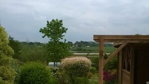 Een verhard terras met een houten pergola, omgeven door weelderige begroeiing en uitzicht op verre bomen en velden.