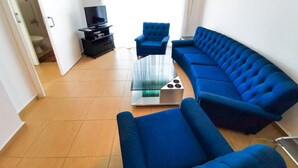 A living room with a blue sofa set, a glass coffee table, and a television on a black stand. - Off Course Seaview Apartment (Larnaca)