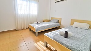 A clean, well-lit room with two single beds, a small bedside table, a nightstand, and a ceiling-mounted air conditioner. - Off Course Seaview Apartment (Larnaca)