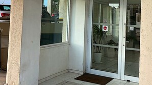 A building entrance with a glass door, a small outdoor area with tiled flooring, and a potted plant inside. - Off Course Seaview Apartment (Larnaca)