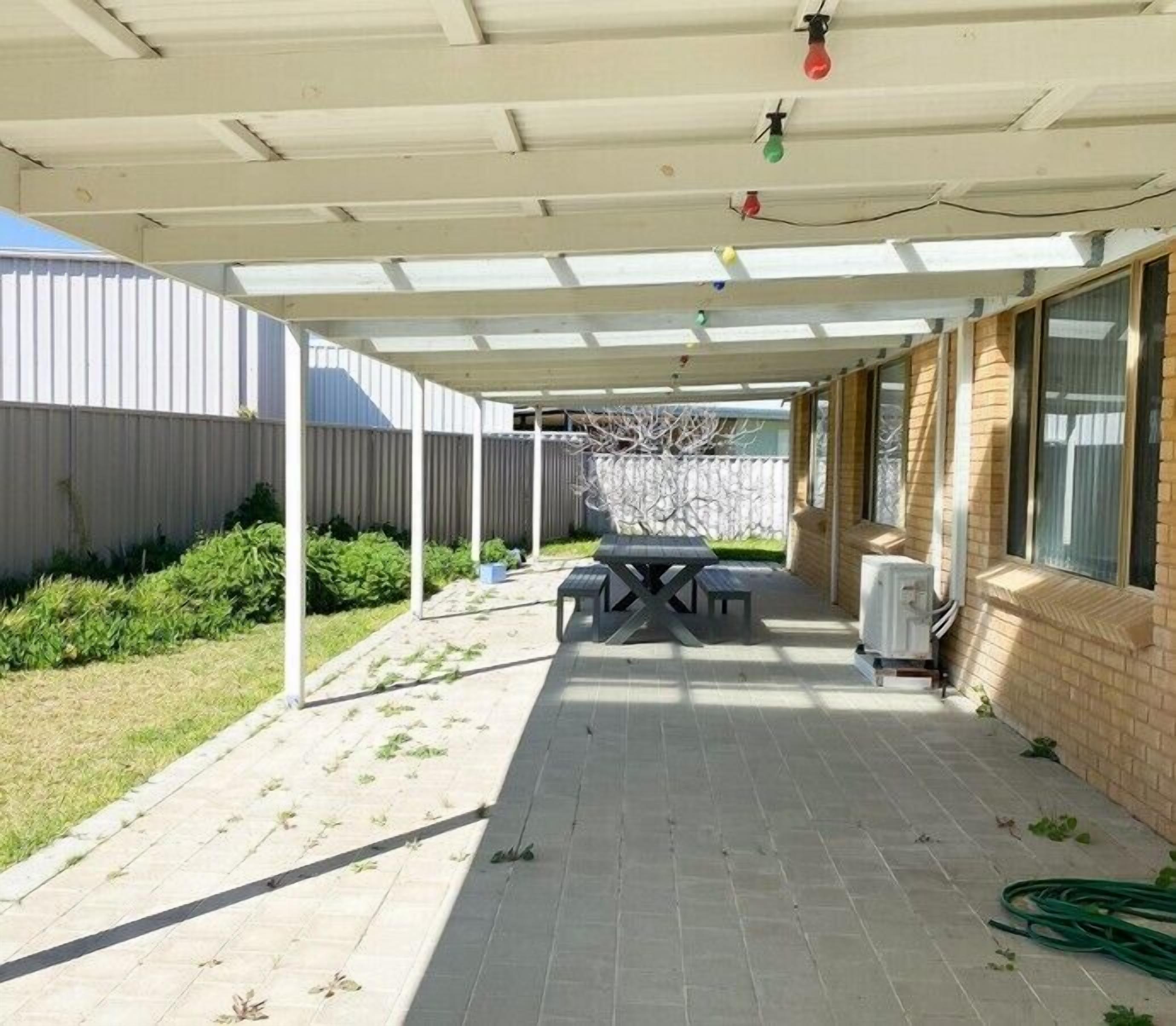 Ein überdachter Patio-Bereich mit Tisch und Stühlen, einem Warmwasserbereiter und einem Garten mit Pflanzen und einem Zaun.
