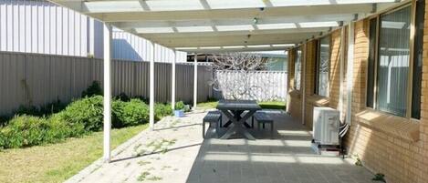 Ein überdachter Patio-Bereich mit Tisch und Stühlen, einem Warmwasserbereiter und einem Garten mit Pflanzen und einem Zaun.