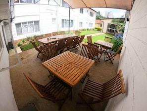 An outdoor patio with a wooden table and chairs, surrounded by a white wall and a garden. - Stay.Plus Hurlingham B&B Suite (Nairobi)