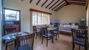 A dining area with wooden chairs and tables, a kitchen area with appliances, and a window with curtains. - Stay.Plus Hurlingham B&B Suite (Nairobi)