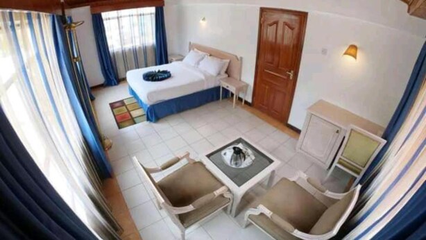 A hotel room with a bed, a desk, two chairs, a small table, a wardrobe, and a window with curtains. - Stay.Plus Hurlingham B&B Suite (Nairobi)