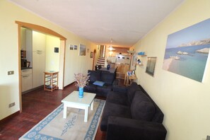 A living room with a sofa, a coffee table, and a rug. There is a kitchen in the background with a refrigerator and a stove.