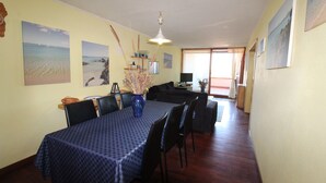 A dining area with a table set for four, a blue patterned tablecloth, and a vase with flowers. There are two framed pictures on the wall, a ceiling fan, and a television in the background. - Tanca Manna three bedrooms apartment 3 bedrooms 2 bathrooms large sea view (Cannigione)