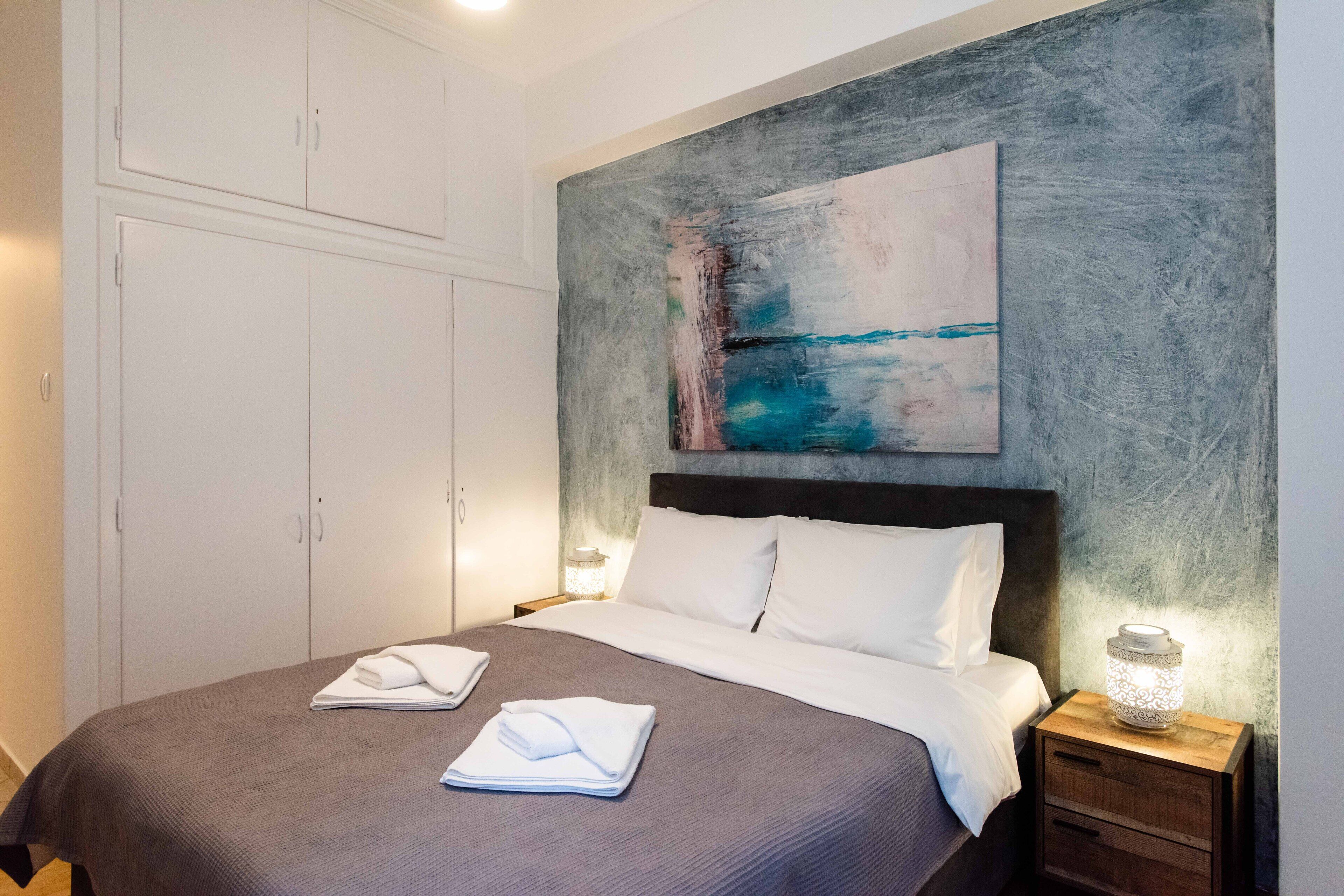 A neatly made bed with white linens, a gray headboard, and a painting above it.