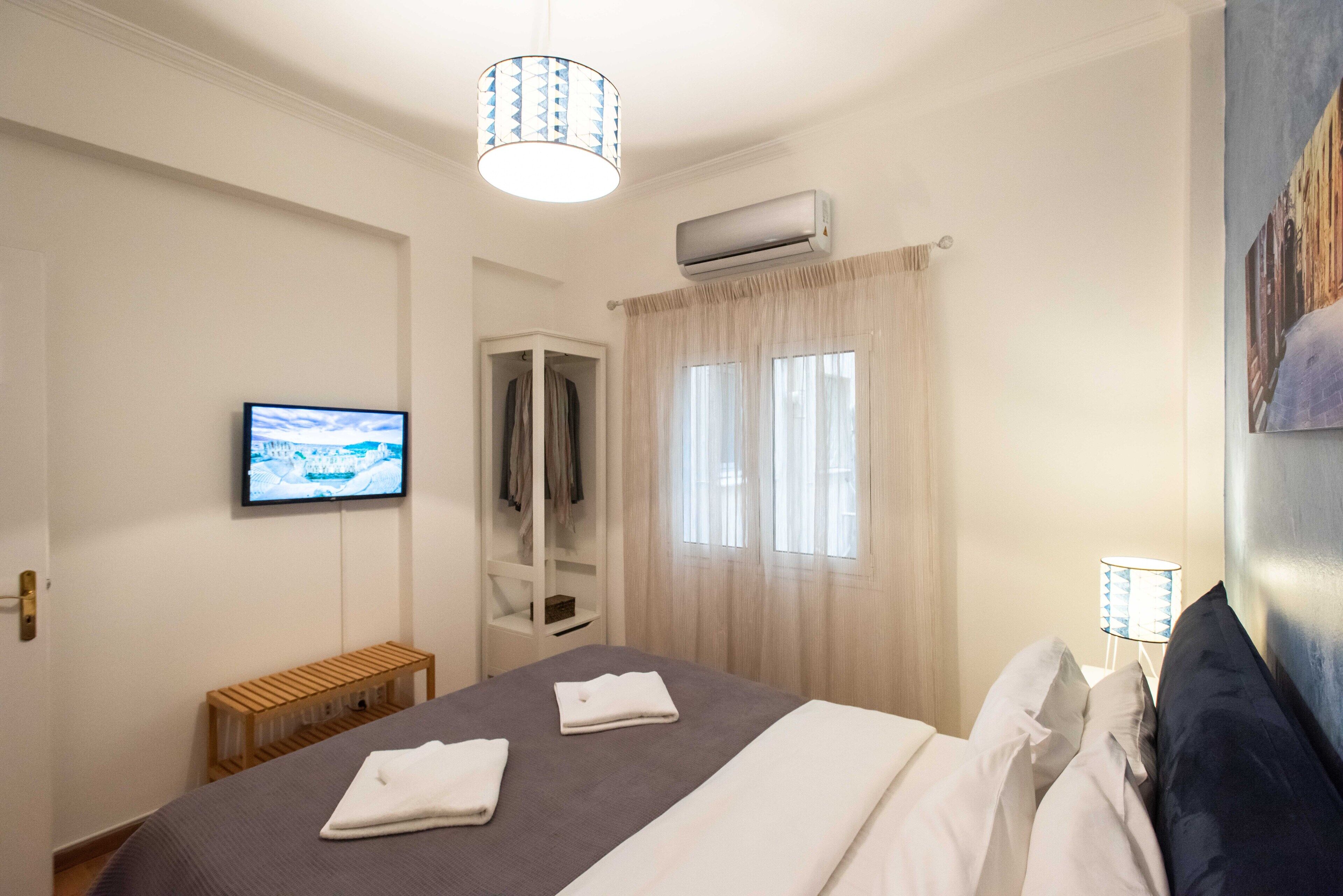 Habitación de hotel con cama, televisor, aire acondicionado, ventana con cortinas, lámpara de noche y un banco.