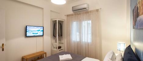 A hotel room with a bed, a television, an air conditioning unit, a window with curtains, a bedside lamp, and a bench.
