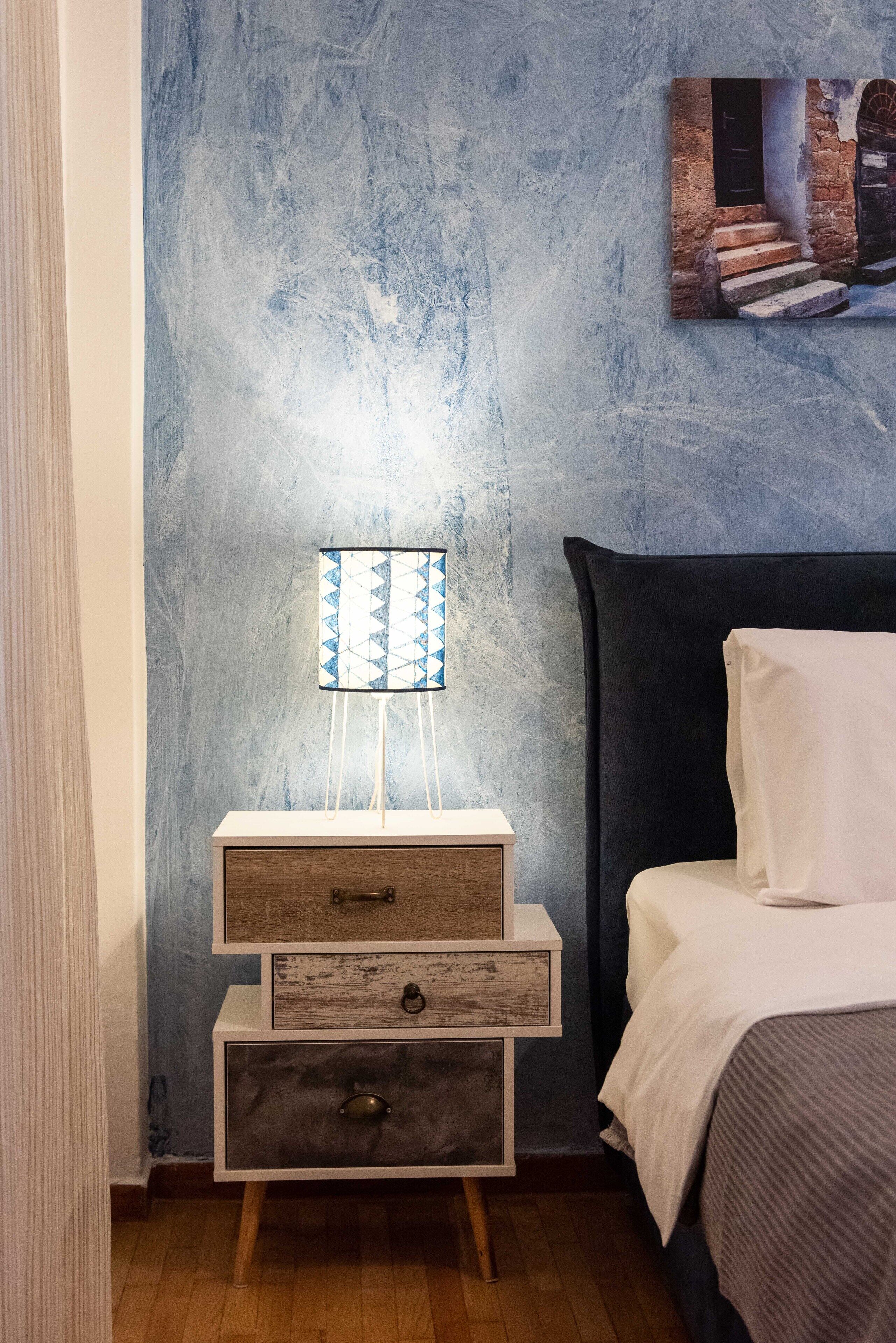A bedroom with a bed, a nightstand with drawers, a lamp, and a textured wall.