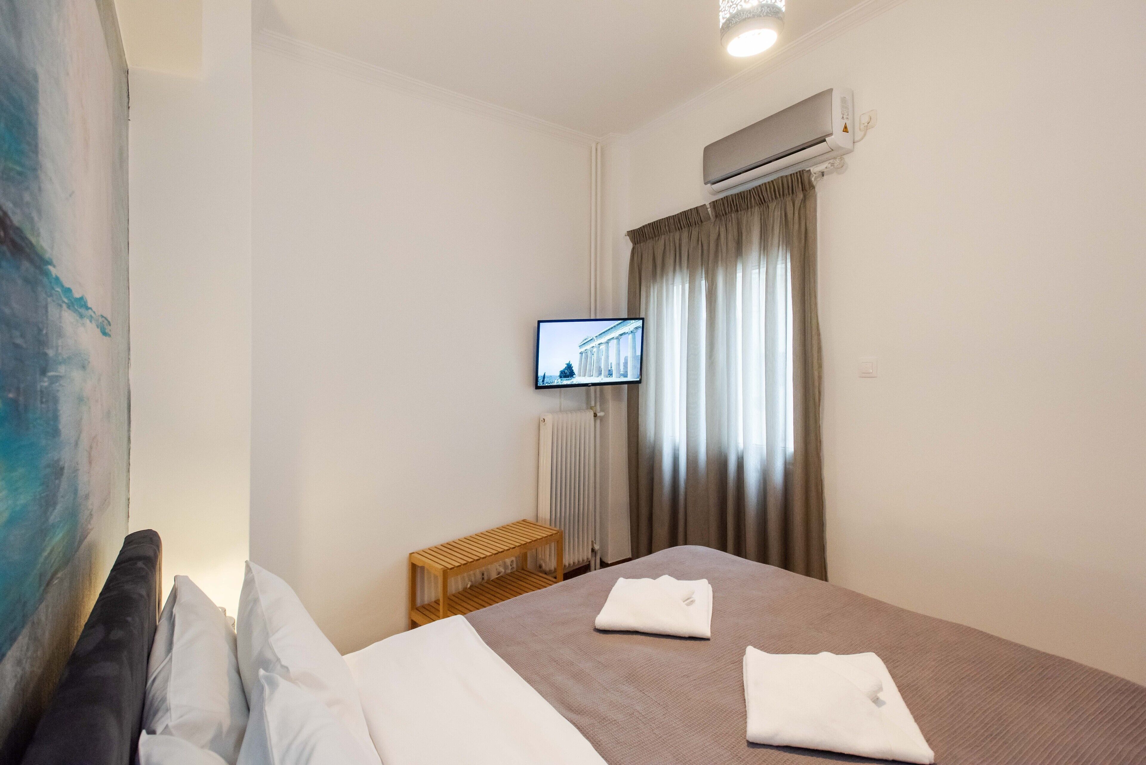 A hotel room with a bed, a TV mounted on the wall, a window with curtains, and an air conditioning unit.