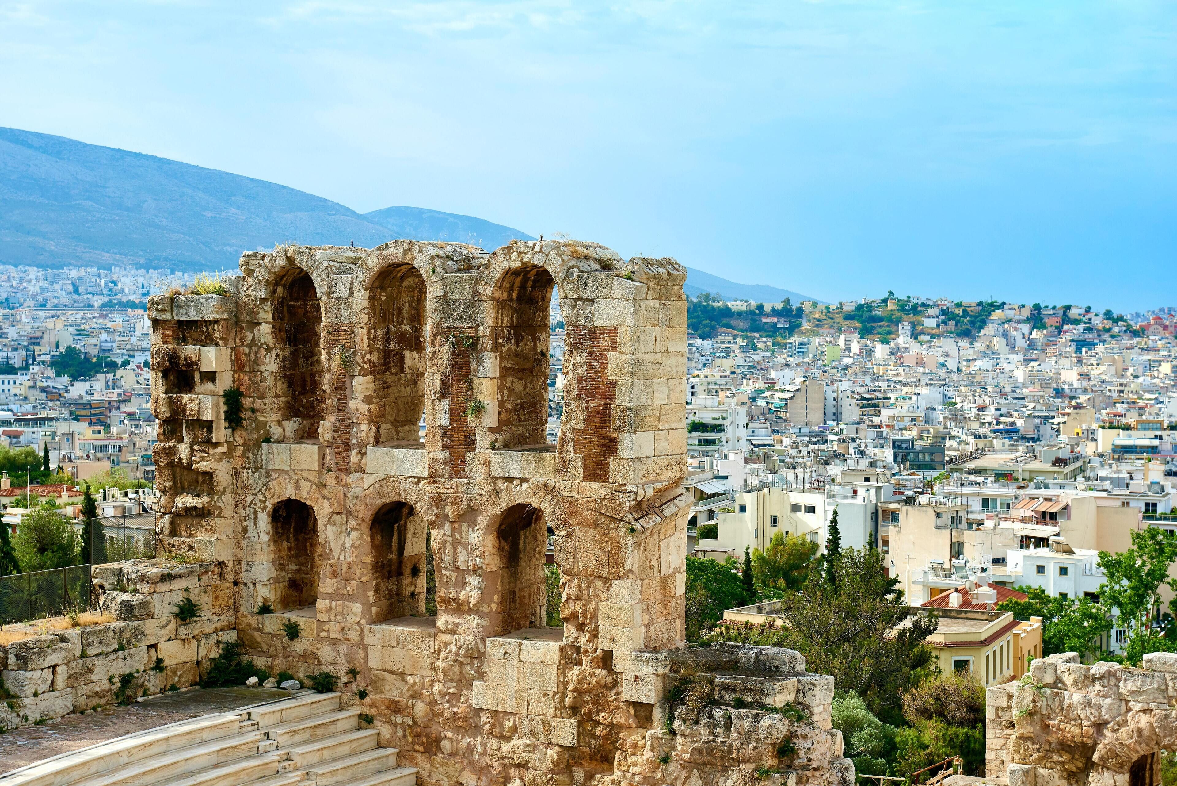 An ancient stone structure with arched openings, surrounded by a cityscape with buildings and a mountain in the background.