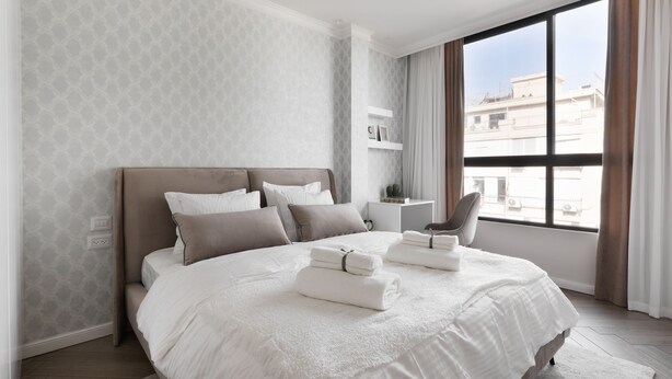 A modern hotel room with a large bed, a ceiling fan, a window with curtains, and a view of the cityscape. - Modern and Central 2BD Apartment in TLV (Tel Aviv)