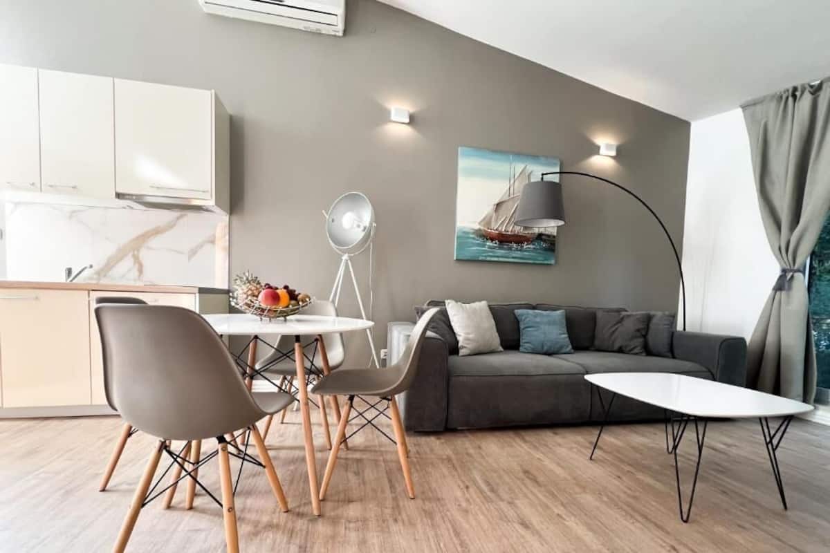 Ein modernes Wohnzimmer mit grauem Sofa, einem weißen Couchtisch und einem Essbereich mit weißem Tisch und Stühlen. An der Wand hängt ein Gemälde eines Segelboots, und ein Stehlampen ist sichtbar.