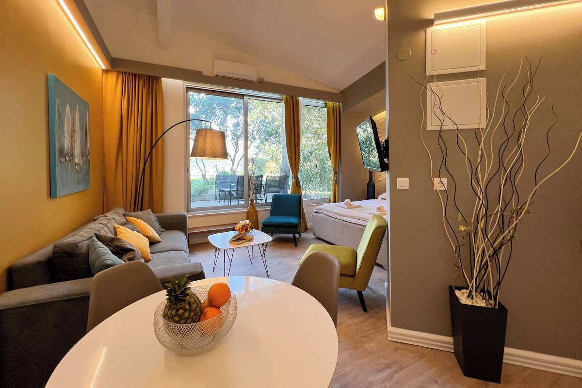 Ein modernes Wohnzimmer mit einer Couch, einem Esstisch mit Obst, einer Lampe und Blick ins Freie.