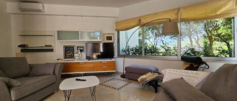 A living room with a sofa, a coffee table, a TV, and a window with a view of trees.