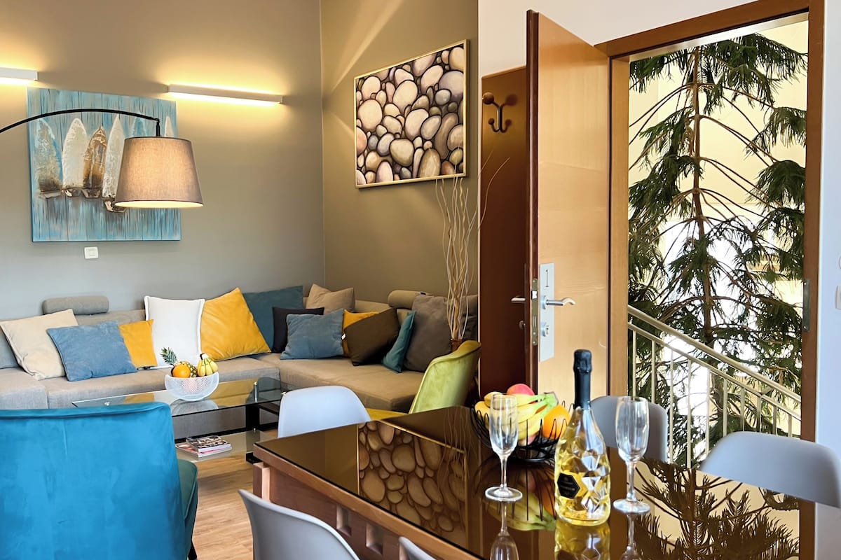Ein modernes Wohnzimmer mit einer Couch, einem Couchtisch und Sesseln. Ein Esstisch mit Weingläsern und einer Obstschale steht bereit. Ein Bild hängt an der Wand, und eine Tür führt auf einen Balkon mit Blick auf Bäume.