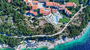 An aerial view of a coastal resort with multiple buildings, a central swimming pool, and a beach area. - Adriatic Resort Apartments (Dubrovnik)