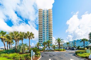 A tall hotel with a central entrance, surrounded by palm trees and a parking area.
