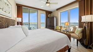 Habitación de hotel con una cama grande, ventilador de techo, vista al océano, balcón y zona de estar con lámpara.