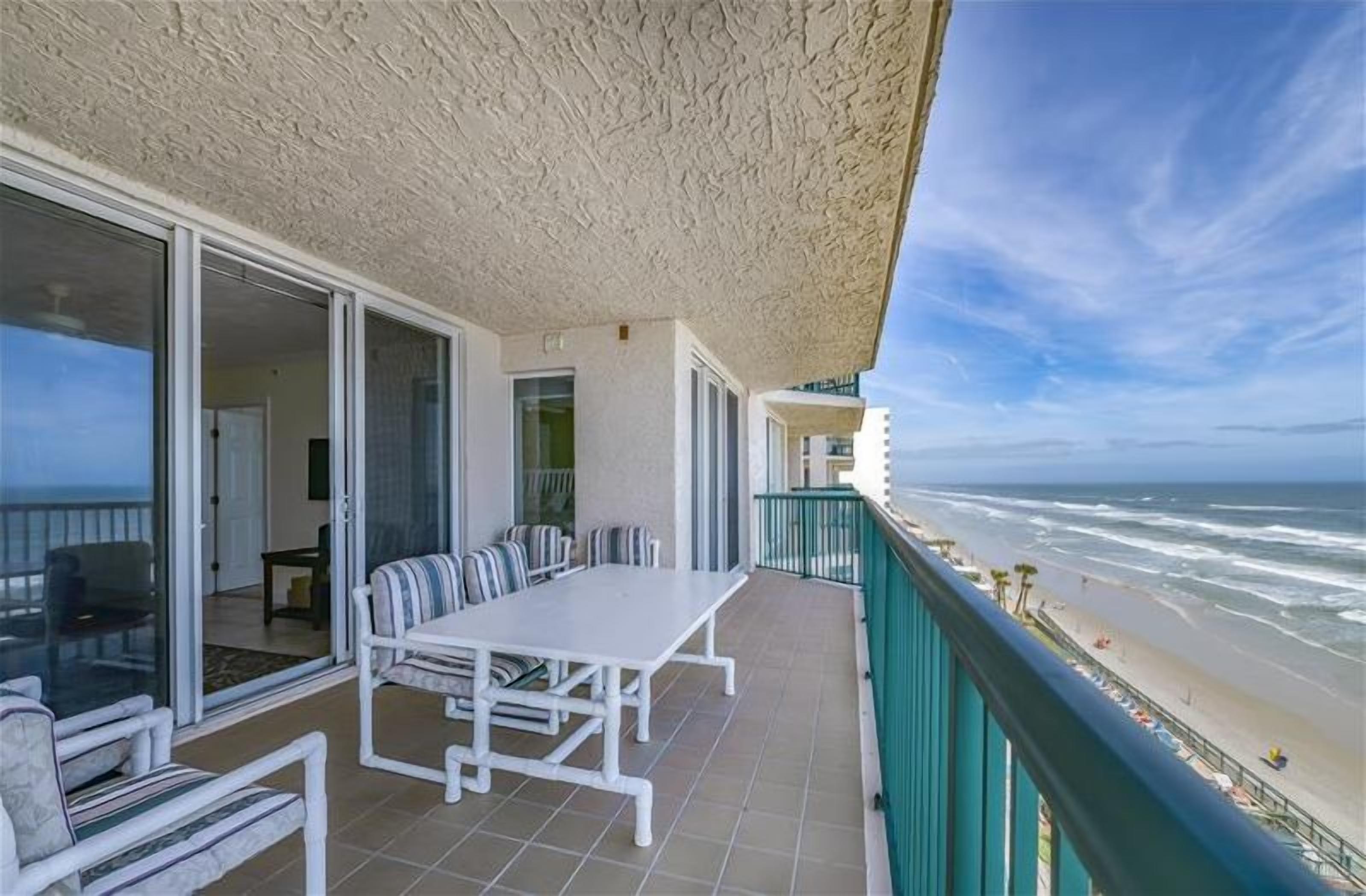 A balcony with a white table and chairs, a glass door leading to a room, and a view of the beach and ocean.