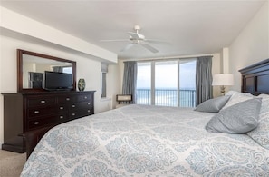 A bedroom with a large bed, a ceiling fan, a dresser, a television, and a view of the ocean.