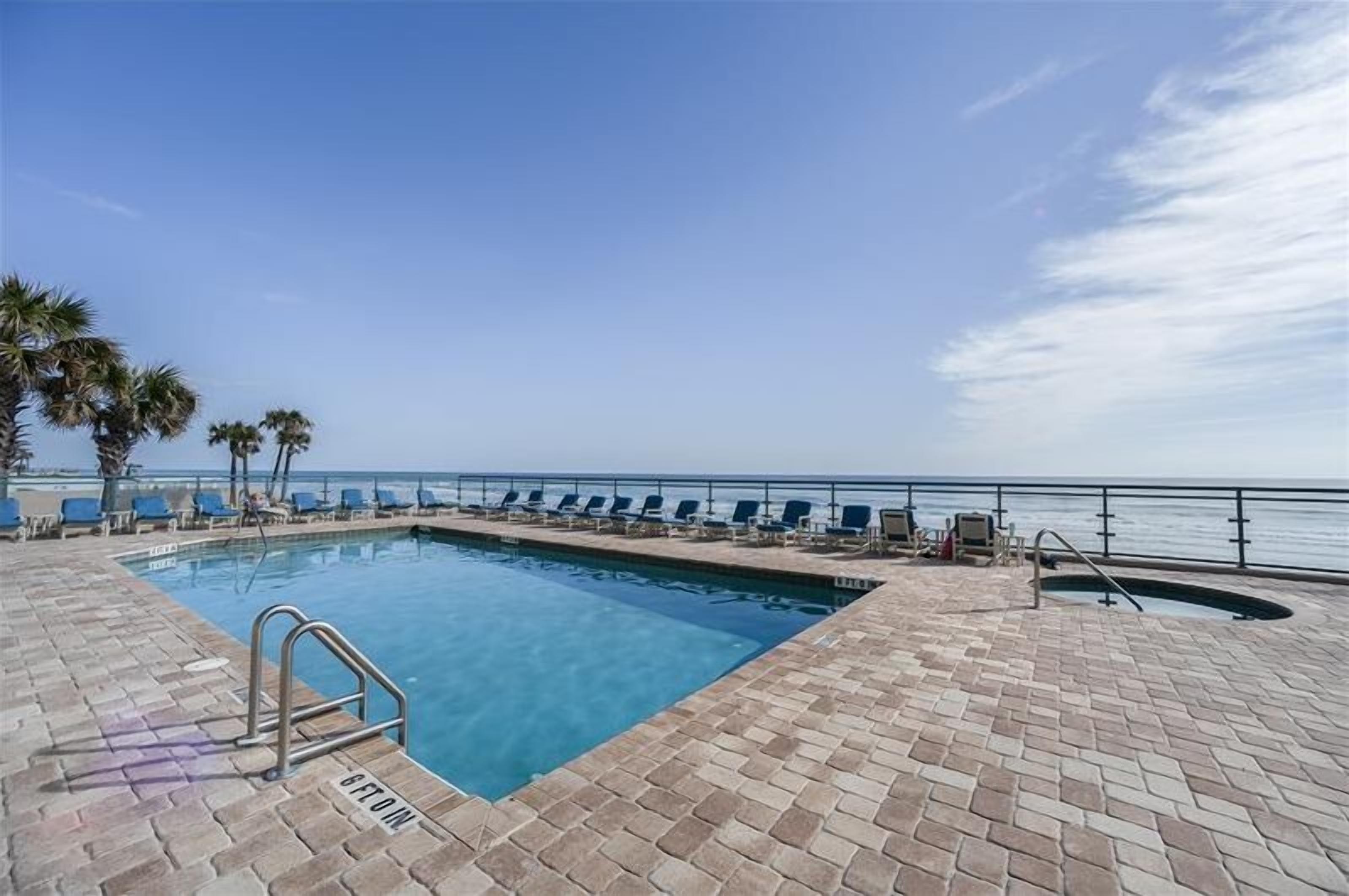 A pool area with a clear view of the ocean, lounge chairs, and palm trees.