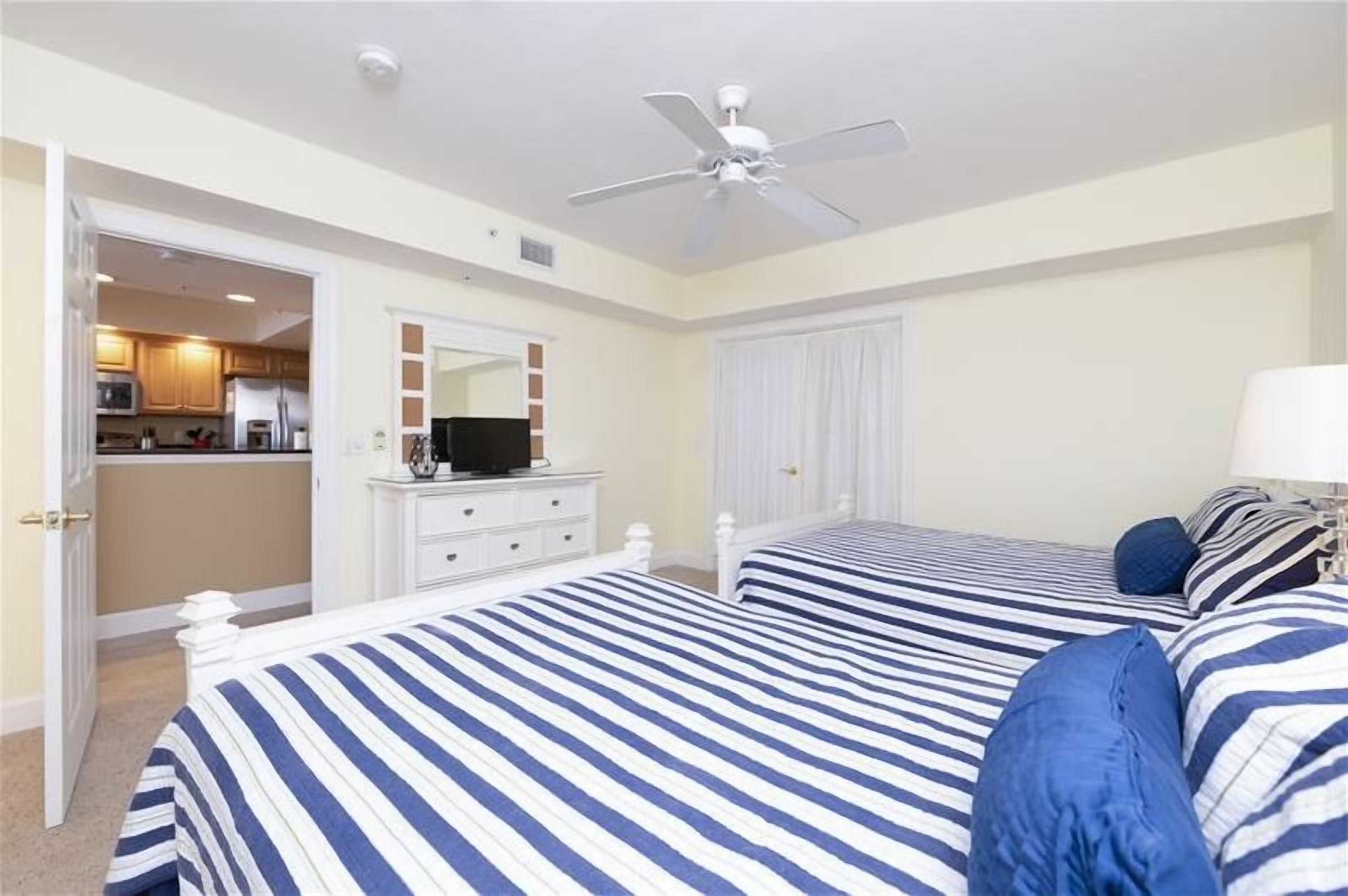 A bedroom with two beds, a ceiling fan, a TV on a dresser, and a view into the kitchen.