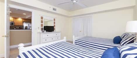A bedroom with two beds, a ceiling fan, a TV on a dresser, and a view into the kitchen.